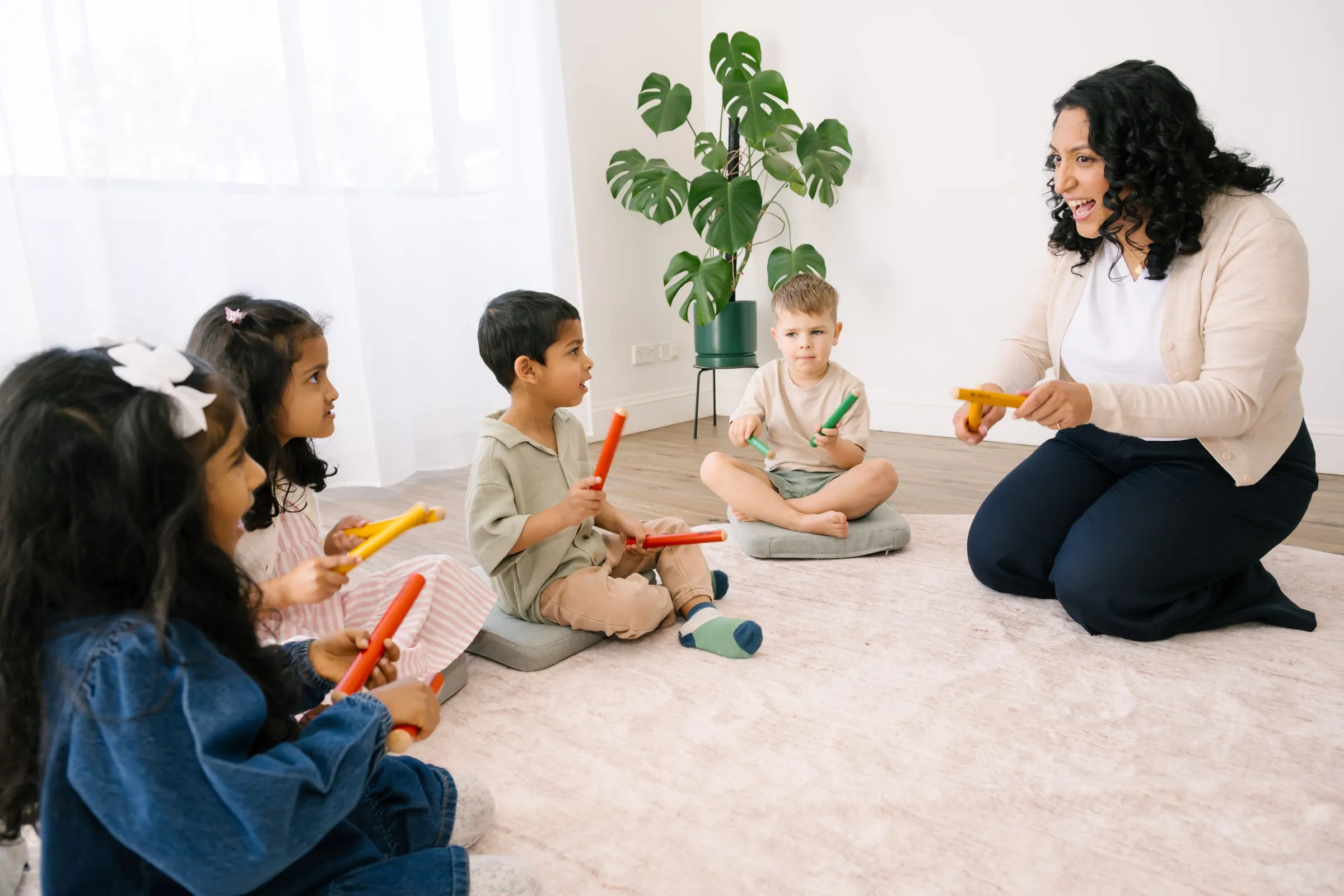Preschoolers playing rhythm sticks with Maria on the floor